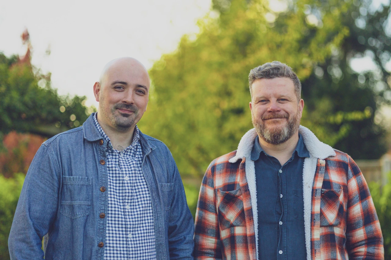 Greg and Biff standing outdoors in front of greenery; the man on the left is Greg - he is bald and wearing a blue denim jacket over a checked shirt; the man on the right is Biff - he has short hair and a beard, wearing a plaid fleece-lined jacket over a dark shirt; both are smiling because they're so happy to be building TrackShift!
